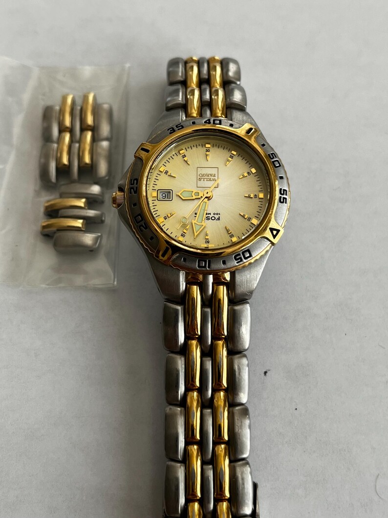 May include: A silver and gold-toned wristwatch with a cream-colored face. The watch has a date display at the 9 o'clock position and a gold bezel. Additional watch links are in a plastic bag.