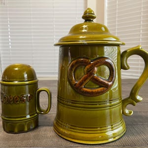 May include: A green ceramic beer stein with a pretzel design and a matching salt shaker with the word "cheese". The stein has a handle and a lid. The salt shaker also has a handle.