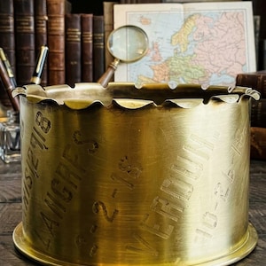 May include: A cylindrical brass shell casing with a scalloped top edge. The exterior surface features engraved text including the words LANGRES and VERDUN, along with dates such as 8-2-18 and 10-26-18, showing a polished metallic finish.