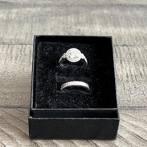 May include: Two silver rings displayed in a black box with a velvet interior. One ring features a large, round, encrusted design, while the other is a simple band with small embellishments. The box sits on a gray wooden surface.
