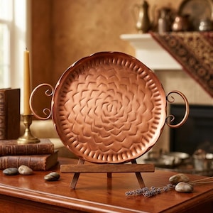 May include: A copper-colored, round decorative tray with a floral pattern in the center and handles. The tray is displayed on a small wooden easel. The tray is approximately 12 inches in diameter.