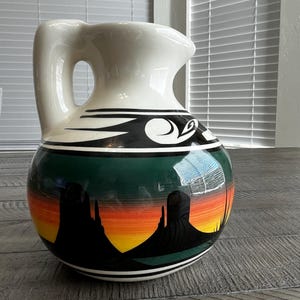 May include: A ceramic pitcher with a white top and handle, featuring a Southwestern desert landscape design. The body of the pitcher showcases a sunset scene with black silhouettes of mesas and a green and black border.