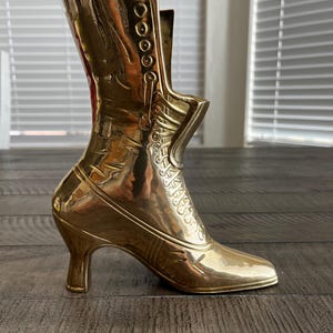 May include: A vintage brass boot-shaped vase. The boot features a high heel, button details, and decorative patterns. The vase is a shiny gold color and reflects the surrounding light. The boot is approximately 7 inches tall.