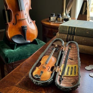 May include: A miniature violin in a decorative case, with a full-sized violin in the background. The miniature violin is displayed in an open case with a bow. The case and violin are brown and gold.
