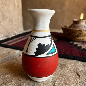 May include: A small, hand-painted ceramic vase with a white neck and a red base. The vase features a black and white geometric design with a turquoise accent. A thin gold band encircles the neck. The vase is displayed on a stone surface.