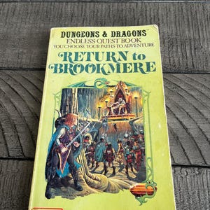 May include: A vintage "Dungeons & Dragons" book titled "Return to Brookmere." The cover features a fantasy illustration of a warrior and a group of figures in a castle setting. The book is yellow with green and orange accents.
