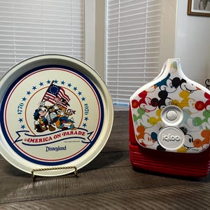 May include: A round metal tray with a white background, blue and red trim, and the text "America on Parade Disneyland." The tray features Mickey Mouse, Donald Duck, and Goofy with an American flag. A red and white Igloo cooler is also in the image.