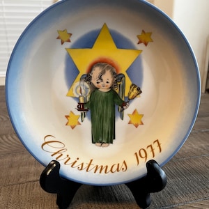 May include: A decorative plate with a blue rim and a central image of an angel holding a candle and a bell, set against a yellow star and surrounded by smaller stars. The plate is inscribed with "Christmas 1977" in gold lettering.