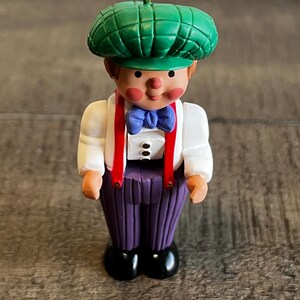 May include: A decorative figurine of a young boy wearing a green cap, white shirt, purple pants, and black shoes. The boy has red suspenders and a blue bow tie. The figurine has a gold loop for hanging.