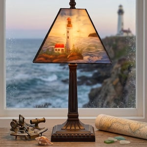 May include: A table lamp with a dark bronze-toned base and a trapezoidal stained glass shade featuring a painted lighthouse scene. The shade displays a white lighthouse with a red top, a small red-roofed building, and ocean waves against a soft sky.