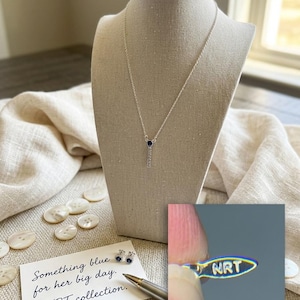 May include: A silver necklace with a small blue gemstone pendant and a matching pair of stud earrings. The necklace is displayed on a mannequin. A card reads "Something blue for her big day. NRT collection."