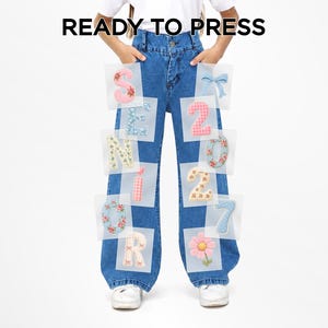 May include: Medium wash blue denim jeans featuring a series of iron-on patches arranged on the legs. The patches display the text SENIOR 2027, a light blue bow, and a pink flower. The text READY TO PRESS is positioned above the jeans.