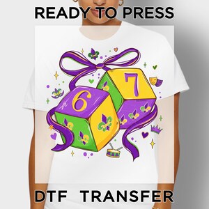 May include: White t-shirt featuring a colorful Mardi Gras design. The graphic includes two dice with the numbers 6 and 7, a purple ribbon, and festive embellishments. Text at the top reads "READY TO PRESS" and at the bottom "DTF TRANSFER".