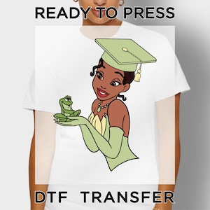 May include: A white t-shirt featuring a graphic of a character wearing a green graduation cap and a green dress, holding a small green frog. The text READY TO PRESS DTF TRANSFER is printed in black at the top and bottom of the image.