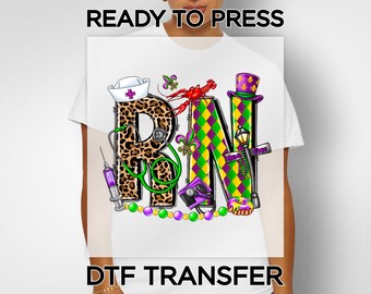 May include: White t-shirt with a colorful graphic featuring the letters "RN" in a Mardi Gras theme. The "R" is leopard print, and the design includes a nurse's hat, stethoscope, syringe, and beads. Text at the top reads "READY TO PRESS" and at the bottom "DTF TRANSFER".