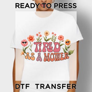 May include: White t-shirt with the text "TIRED AS A MOTHER" in a retro font, surrounded by cartoon flowers with sunglasses. The words "READY TO PRESS" and "DTF TRANSFER" are printed above and below the design.