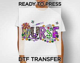 May include: White t-shirt with a colorful "NURSE" design. The design includes a nurse's cap, stethoscope, syringe, and thermometer. The word "NURSE" is in various patterns and colors, with the text "READY TO PRESS" and "DTF TRANSFER" above and below the design.