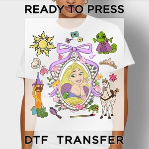 May include: A white t-shirt featuring a DTF transfer with a central oval portrait of a blonde character in a pearl frame, topped with a purple bow. Surrounding elements include a sun, a tower, a horse, a chameleon, a brush, and flowers.