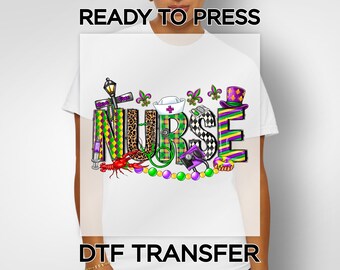 May include: White t-shirt featuring a colorful graphic design with the word "NURSE" in large, decorative letters. The design incorporates medical symbols, a crawfish, and Mardi Gras elements. The text "READY TO PRESS" and "DTF TRANSFER" are also visible.