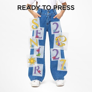 May include: Blue denim jeans featuring heat transfer decals with the text READY TO PRESS at the top. The designs include the word SENIOR in yellow and purple letters, the number 2027, a cartoon character with long blonde hair, and a sun icon.