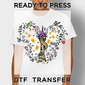 May include: A white t-shirt featuring a graphic print of a tall stone tower with a purple roof and long blonde hair cascading down. The design is framed by floral vines and floating lanterns in a Mickey Mouse ear shape. Text reads READY TO PRESS DTF TRANSFER.