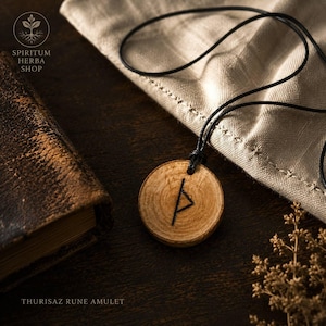 Op de afbeelding: Een ronde houten hanger met een donker gegraveerd Thurisaz-runesymbool, hangend aan een dun zwart koord. De natuurlijke houtnerf van het lichte hout is zichtbaar op het oppervlak van het amulet, dat naast een gestructureerde stof ligt.