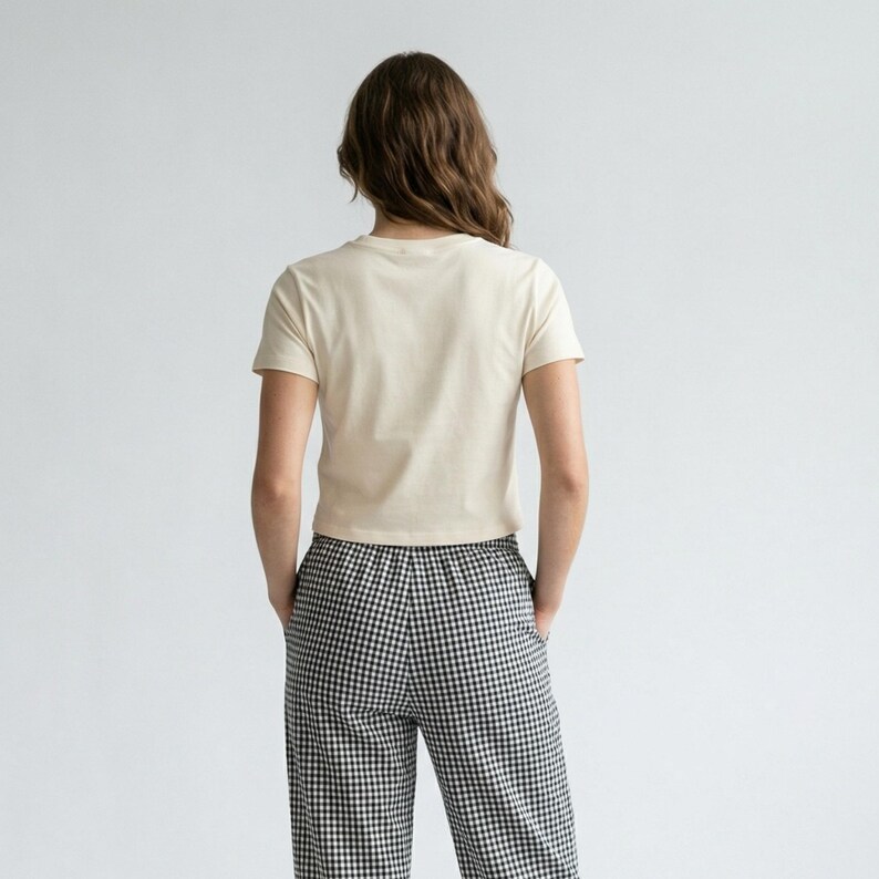 May include: A person wearing a cream-colored, short-sleeved t-shirt and black and white checkered pants. The model is facing away from the camera, with hands in pockets. The background is a plain white.