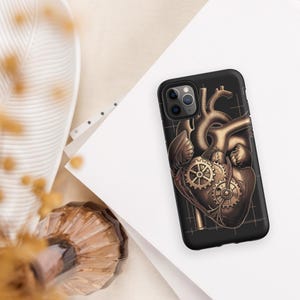 May include: Black phone case featuring a steampunk-style illustration of a mechanical heart. The design incorporates gears and a metallic gold color scheme. The case has a matte finish and a grid pattern background.
