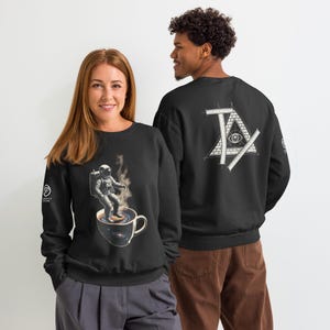 Op de afbeelding: Zwart sweatshirt met een afbeelding van een astronaut die surft op een kop koffie, met opstijgende stoom. De achterkant van het sweatshirt heeft een wit geometrisch ontwerp. Het sweatshirt wordt door twee personen gedragen.