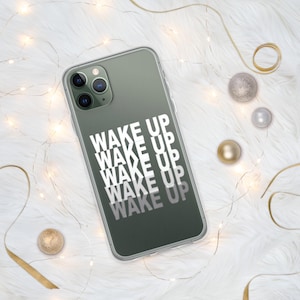 May include: A clear phone case with the words "WAKE UP" stacked vertically in white. The phone case is on a white fluffy surface with gold and silver decorative balls and a gold ribbon.
