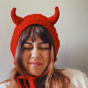 以下が含まれることがあります： 手作りの赤いかぎ針編みの悪魔の角の帽子。帽子には2つの湾曲した角があり、あごの下で結ばれています。ハロウィーンやコスチュームパーティーにぴったりの楽しいアクセサリーです。