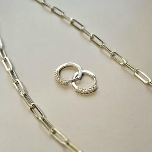 May include: A pair of silver hoop earrings with small, clear gemstones set along the front. The earrings are on a light gray surface, with two silver chain necklaces in the background.