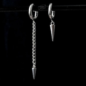 May include: A pair of silver-toned hoop earrings featuring dangling charms. One earring has a long chain ending in a metal spike, while the other features a single, shorter metal spike. The text tender armor is visible in the center.