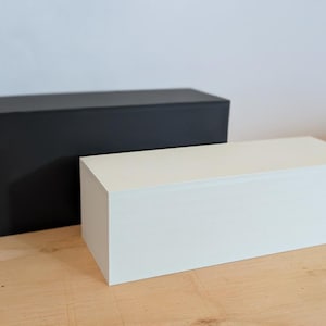 May include: Two rectangular boxes, one black and one white, are positioned on a light wood surface. The black box is larger and stands behind the white box. Both boxes have a matte finish and a simple, modern design.