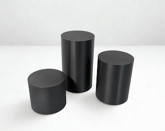 May include: Three black cylindrical objects of varying heights are arranged on a white surface. The cylinders have a matte finish and vertical ridged detailing. The objects are solid and appear to be made of a dense material.