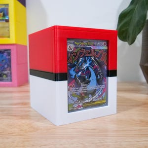 May include: A red, black, and white cube-shaped card holder with a Pokemon card visible in the front. The card features a blue and black dragon-like creature. Other similar holders are stacked in the background.