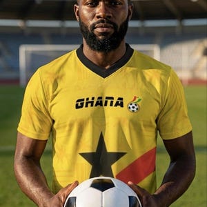 Pu&ograve; includere: Una persona indossa una maglia da calcio gialla del Ghana con una stella nera e la scritta "GHANA". La persona tiene in mano un pallone da calcio bianco e nero. Lo sfondo &egrave; un campo verde e una porta.