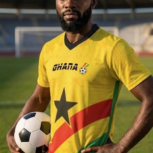 Pu&ograve; includere: Una maglia da calcio gialla con la scritta "GHANA" e una grafica di pallone da calcio. La maglia presenta una stella nera e strisce rosse, gialle e verdi. Un pallone da calcio &egrave; tenuto davanti alla maglia.
