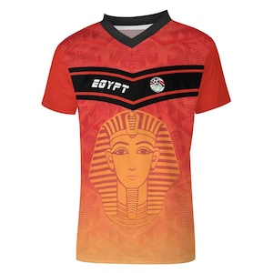 Op de afbeelding: Rood en oranje voetbalshirt met een zwarte V-hals. Het shirt heeft het woord "EGYPT" in het wit, een voetbalembleem en een grote afbeelding van een farao's gezicht. De mouwen zijn effen rood.