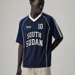 Op de afbeelding: Een marineblauw V-hals shirt met witte biezen, de tekst SOUTH SUDAN in witte blokletters, het nummer 10 en de vlag van Zuid-Soedan. Het shirt heeft witte strepen langs de schouders en wordt gedragen met een lichtbeige broek.
