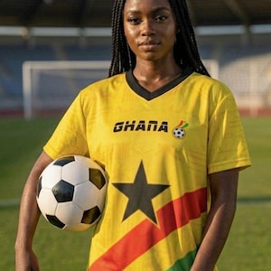Pu&ograve; includere: Una maglia da calcio gialla con la scritta "GHANA" e una grafica di pallone da calcio. La maglia presenta una stella nera e strisce rosse, gialle e verdi. Un pallone da calcio &egrave; tenuto davanti alla maglia.