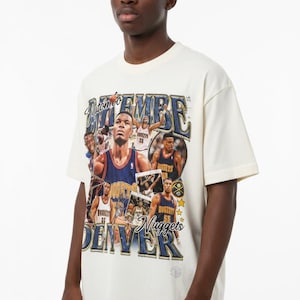 Può includere: T-shirt color crema con un collage di immagini di giocatori di basket e la scritta "Nuggets Denver". Il design include accenti blu e oro, con il nome del giocatore in evidenza.
