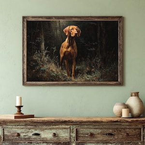 May include: A framed print depicting a Vizsla dog in a forest setting. The dog, with its reddish-brown coat, is the central figure. The artwork is enclosed in a dark wooden frame, with a dark forest as the backdrop.