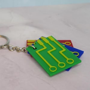 Pode incluir: Um porta-chaves com um anel e corrente prateados segurando três pingentes coloridos em forma de placa de circuito. Os pingentes são verdes, azuis e vermelhos, com desenhos de circuitos amarelos. O porta-chaves está em uma superfície rosa claro.