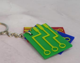 Könnte beinhalten: Ein Schlüsselanhänger mit einem silbernen Ring und einer Kette, die drei bunte Anhänger in Form von Leiterplatten hält. Die Anhänger sind grün, blau und rot, mit gelben Schaltungsdesigns. Der Schlüsselanhänger befindet sich auf einer hellrosa Oberfläche.