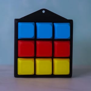 Puede incluir: Un marco cuadrado negro que contiene una cuadrícula de tres por tres con nueve botones cuadrados, lisos y elevados. La fila superior presenta tres botones azules, la fila central tiene tres botones rojos y la fila inferior tres botones amarillos.