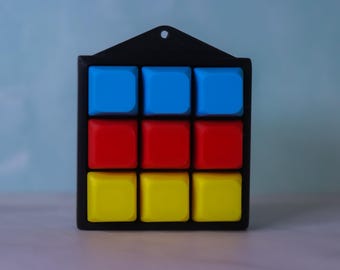 Könnte beinhalten: Ein quadratischer schwarzer Rahmen mit einem drei-mal-drei Raster aus neun erhabenen, glatten, quadratischen Tasten. Die obere Reihe besteht aus drei blauen Tasten, die mittlere aus drei roten und die untere Reihe aus drei gelben Tasten.
