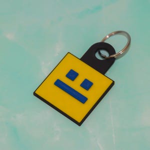 May include: A yellow square keychain with a black border and a black loop. The keychain features a blue horizontal line and two blue squares, resembling a face. A silver key ring is attached.