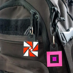 K&ouml;nnte beinhalten: Zwei geometrische Schl&uuml;sselanh&auml;nger h&auml;ngen an einem schwarzen Rucksack. Ein Schl&uuml;sselanh&auml;nger hat ein rot-wei&szlig;-schwarzes Design, w&auml;hrend der andere rosa mit einem lila Quadrat in der Mitte ist. Beide haben schwarze Anh&auml;nger und silberne Schl&uuml;sselringe.