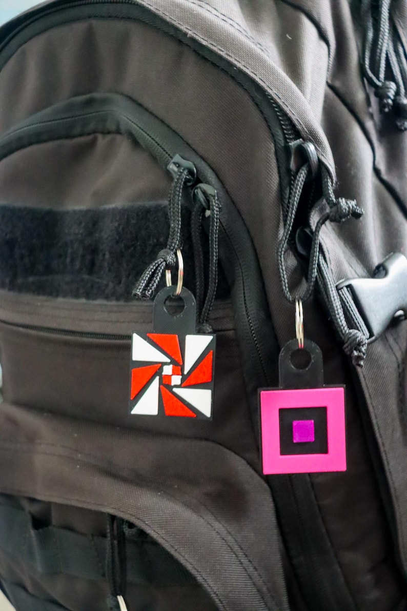 K&ouml;nnte beinhalten: Zwei geometrische Schl&uuml;sselanh&auml;nger h&auml;ngen an einem schwarzen Rucksack. Ein Schl&uuml;sselanh&auml;nger hat ein rot-wei&szlig;-schwarzes Design, w&auml;hrend der andere rosa mit einem lila Quadrat in der Mitte ist. Beide haben schwarze Anh&auml;nger und silberne Schl&uuml;sselringe.